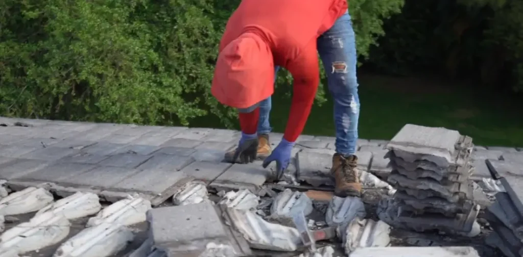 A worker in gloves carefully removes heavy grey concrete tiles from an old roof.